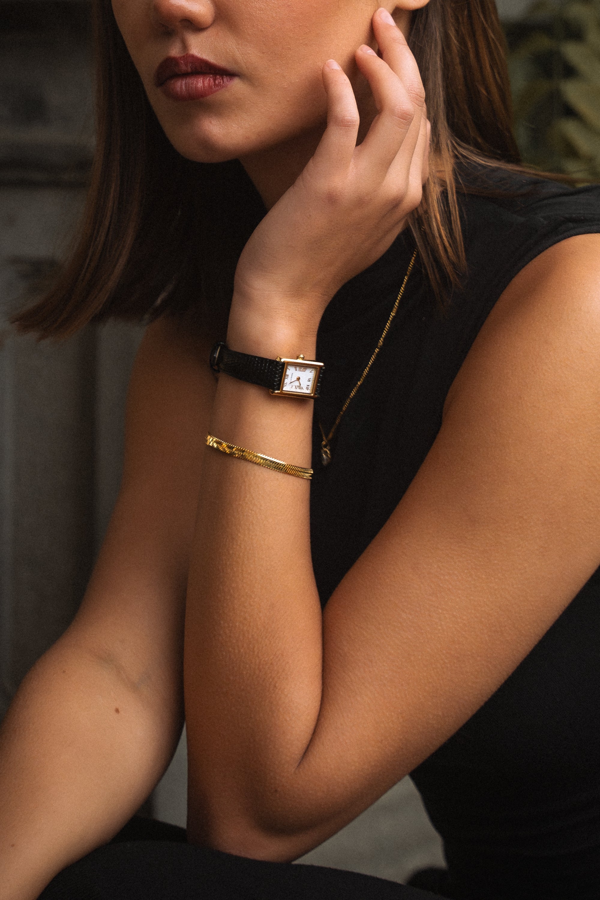 Close-up of the Lucienta Amara gold square watch with black leather strap, styled on a model wearing a black outfit, paired with a gold bracelet.