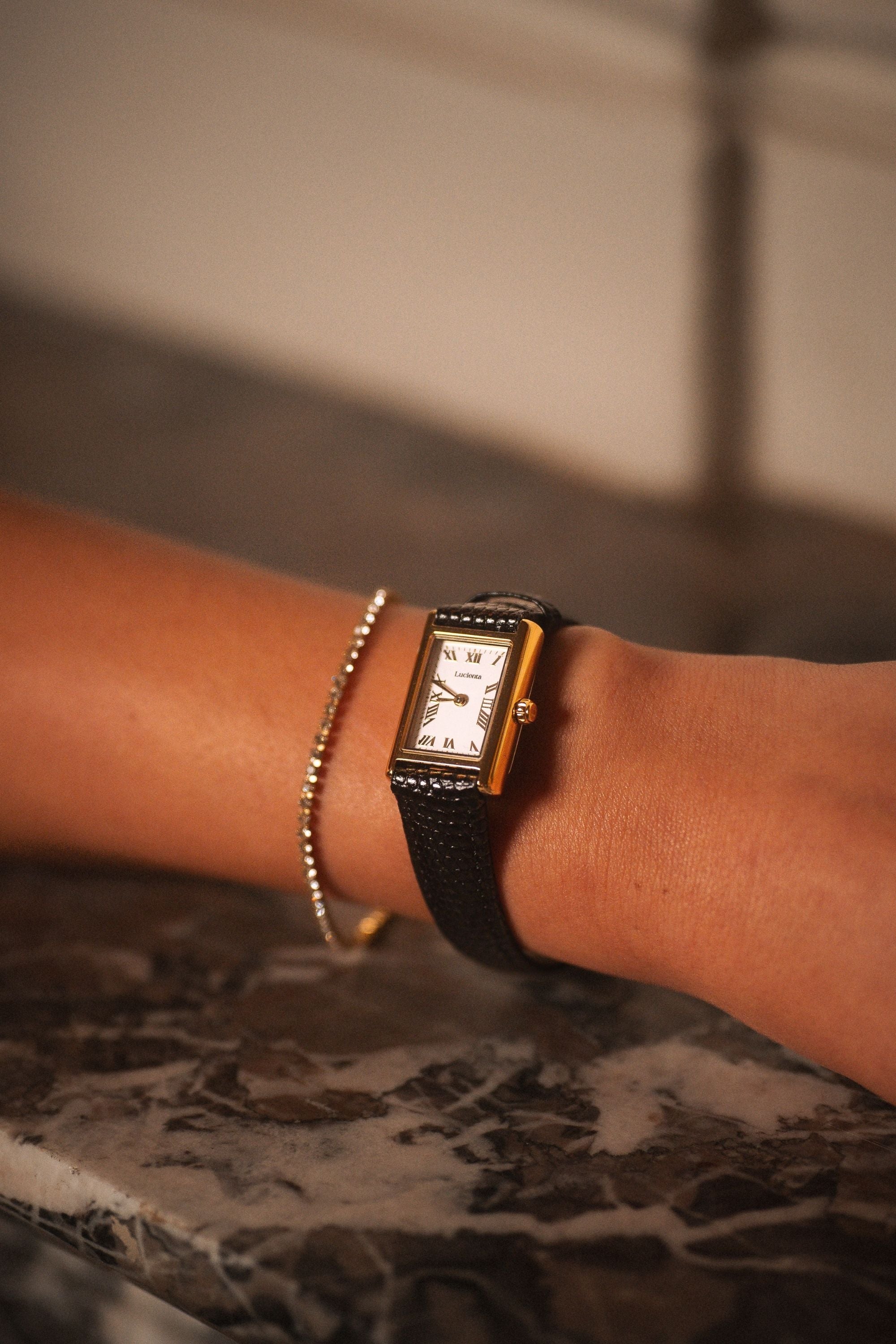 Lucienta Amara gold square watch with black leather strap, styled with a delicate gold bracelet on a marble surface.