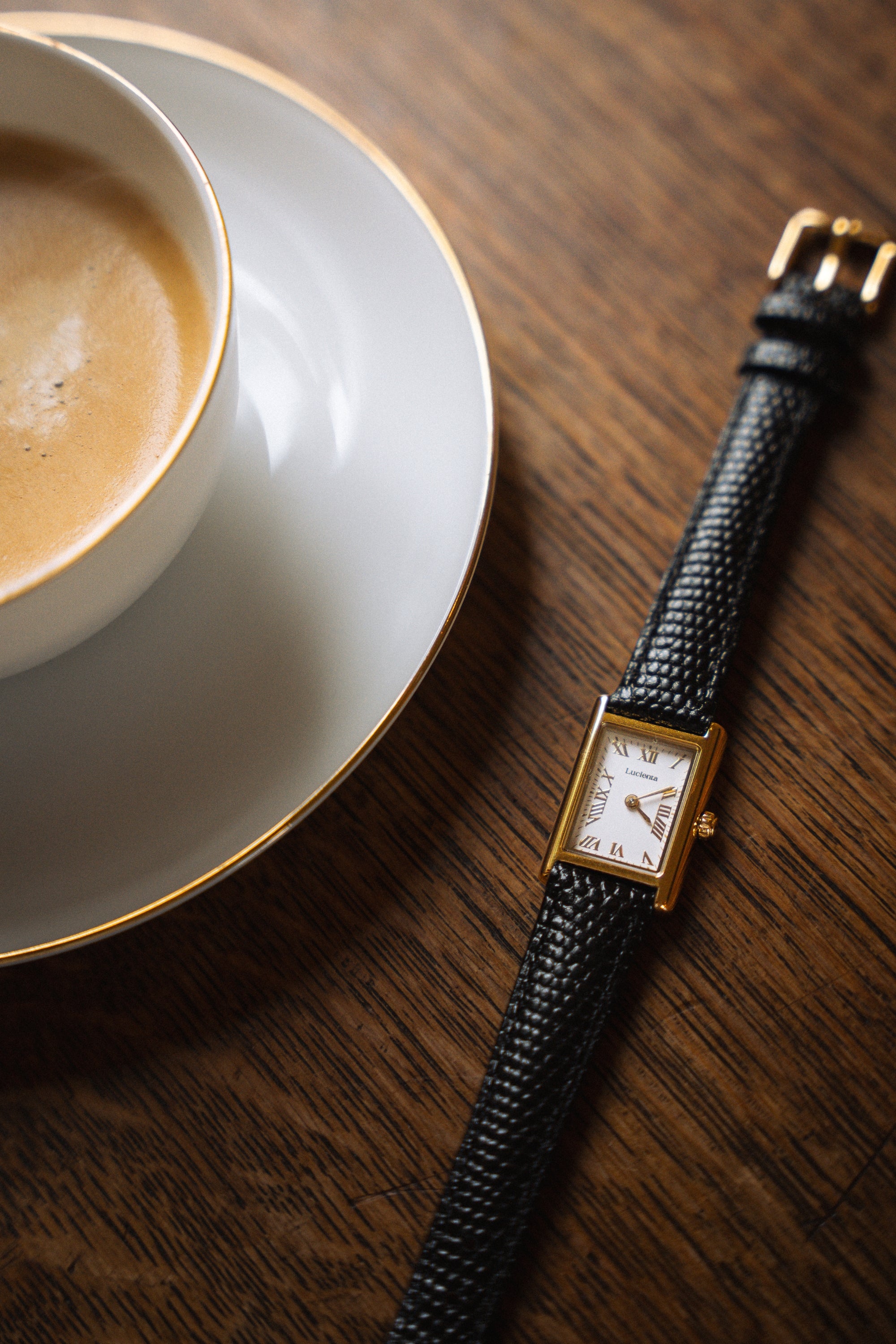 Lucienta Amara gold square watch with black leather strap, elegantly placed beside a cup of coffee on a wooden table with a warm ambiance.